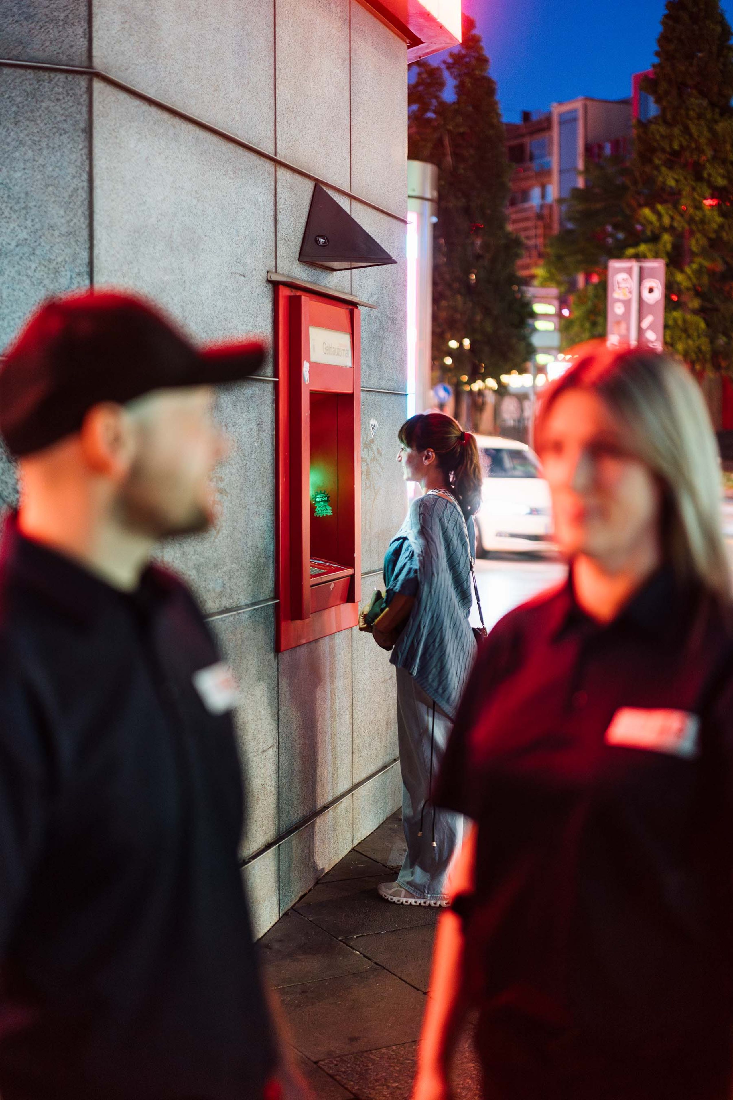 Frau hebt Geld am Automaten ab, HLS-Mitarbeiter sichern den Bereich